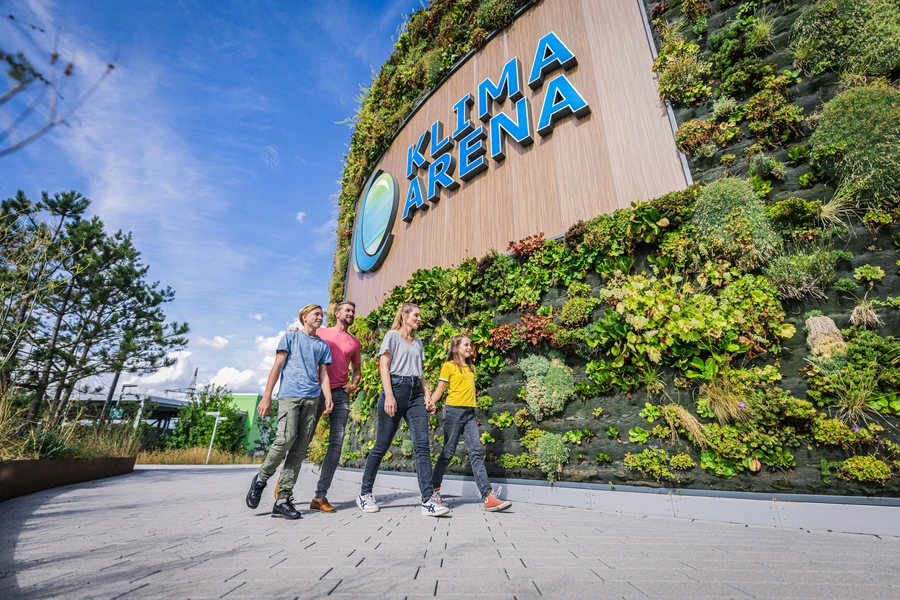 Familie l&auml;uft auf dem Vorplatz der KLIMA ARENA entlang der begr&uuml;nten Fassade mit Logo.